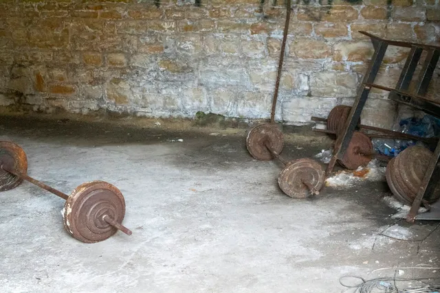 Rusted barbells and loose weight plates left on a bare concrete floor in a disused room
