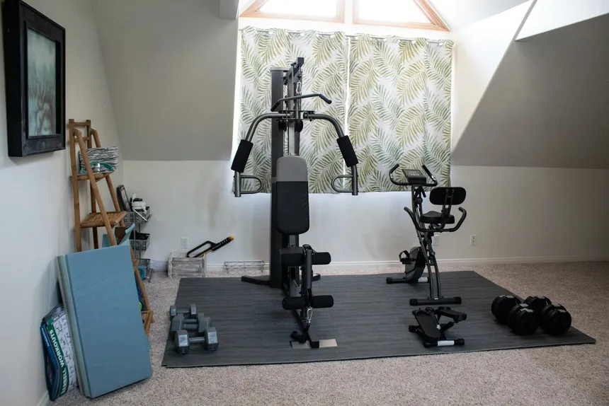 An all-in-one cable machine set up in a small attic room, with a mat, a folded bench and a rack of dumbbells along the wall
