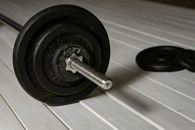 A screw-collar dumbbell bar with the thread and locking collar exposed, plates stacked on the floor beside it
